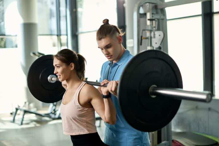 Young athletic woman exercising with barbell with help of personal trainer in a gym.